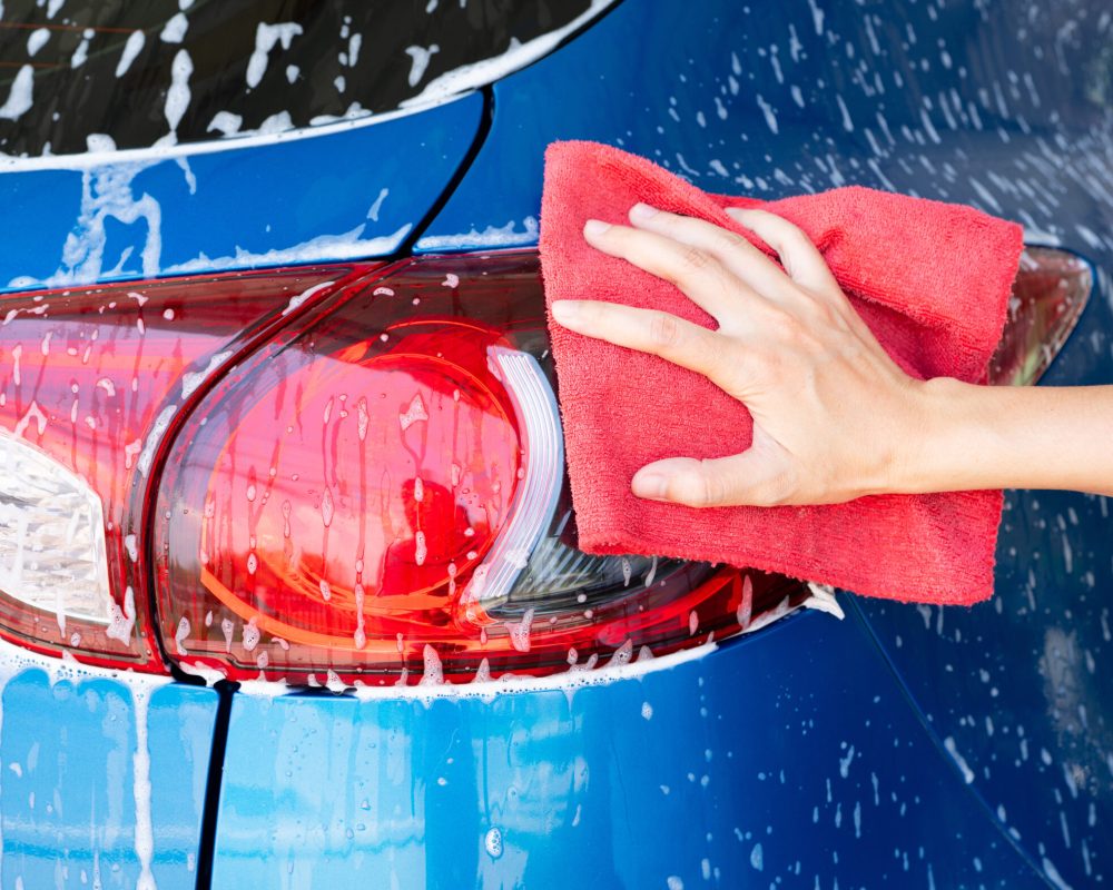 Car wash service. Car covered with white soap foam. Man hand holding red microfiber cloth and polish tail light of blue car. Auto care service business concept. Man cleaning and detailing luxury car.